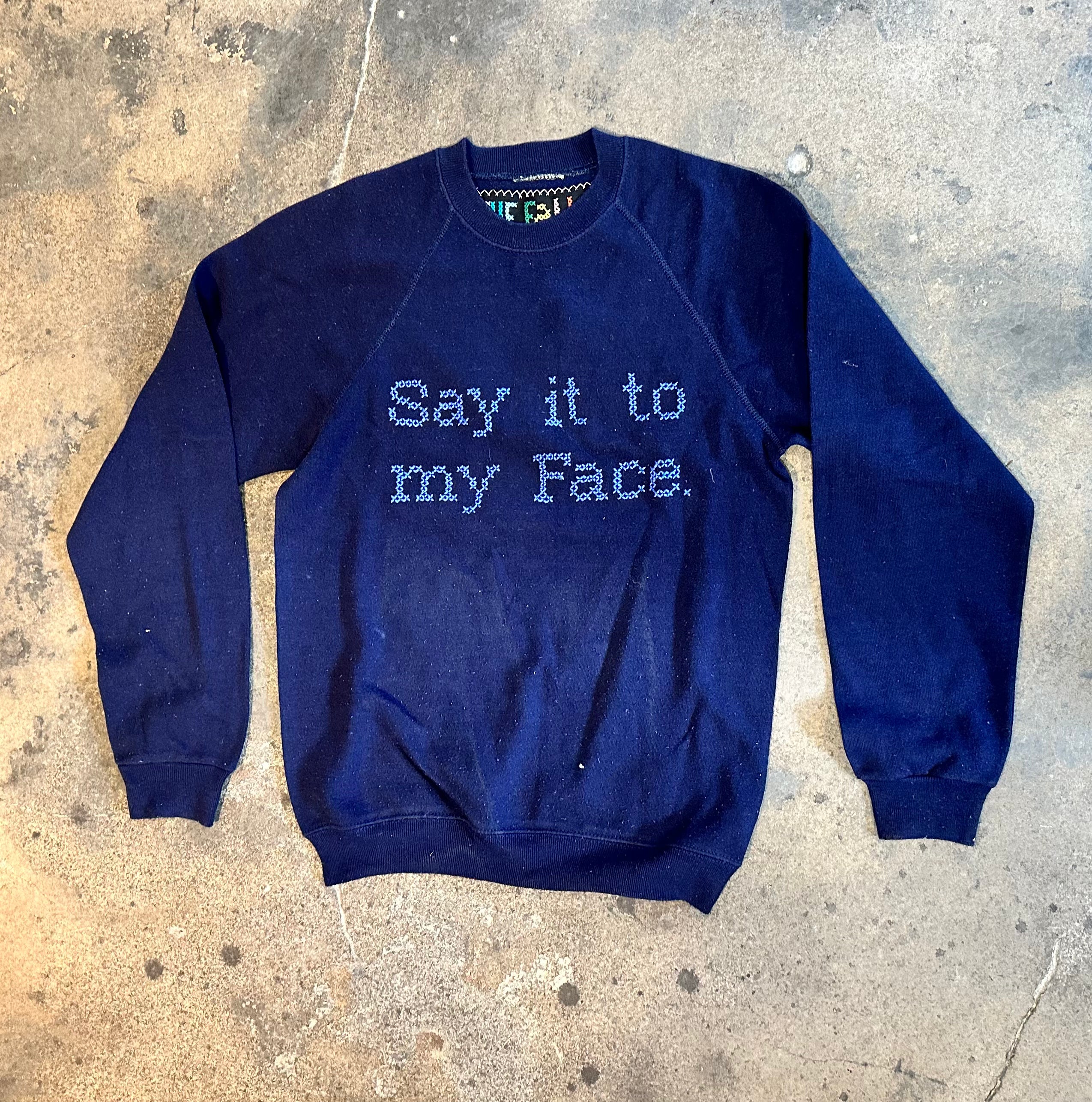 Upcycled sweatshirt with embroidery saying  “Say it to my face”, front view. 